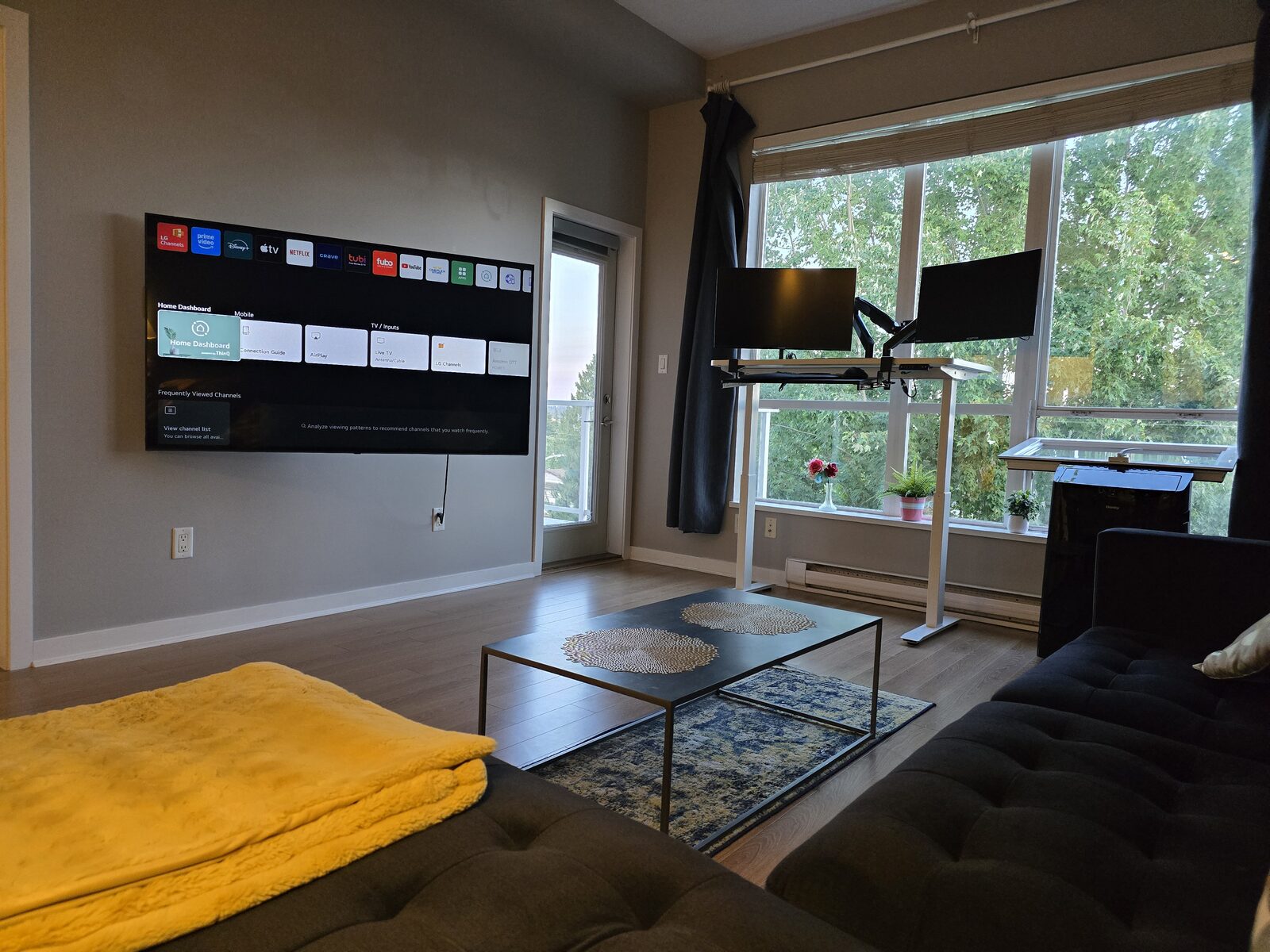 Wide view of the living room showing the sectional sofa, standing desk, and 65 inch smart TV with natural light from floor-to-ceiling windows
